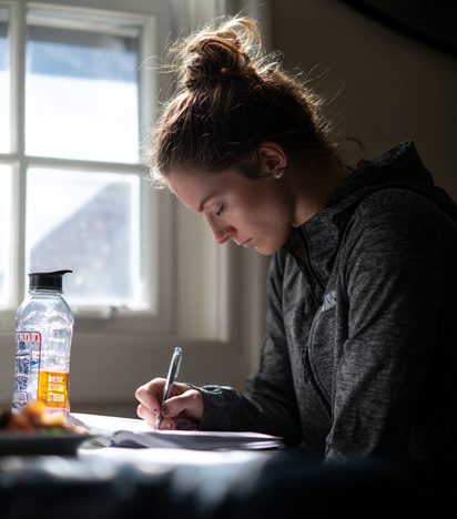 Athlete writing in a nutrition journal beside a water bottle by a window