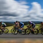 Peloton of cyclists riding at speed across open countryside under a dramatic cloudy sky