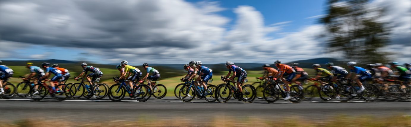 Peloton of cyclists riding at speed across open countryside under a dramatic cloudy sky