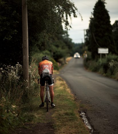 Cyclist riding away down a quiet, hedge-lined country lane in evening light